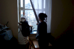 A rejected asylum seeker looks out the window of her darkened room with a refugee camp worker standing nearby.