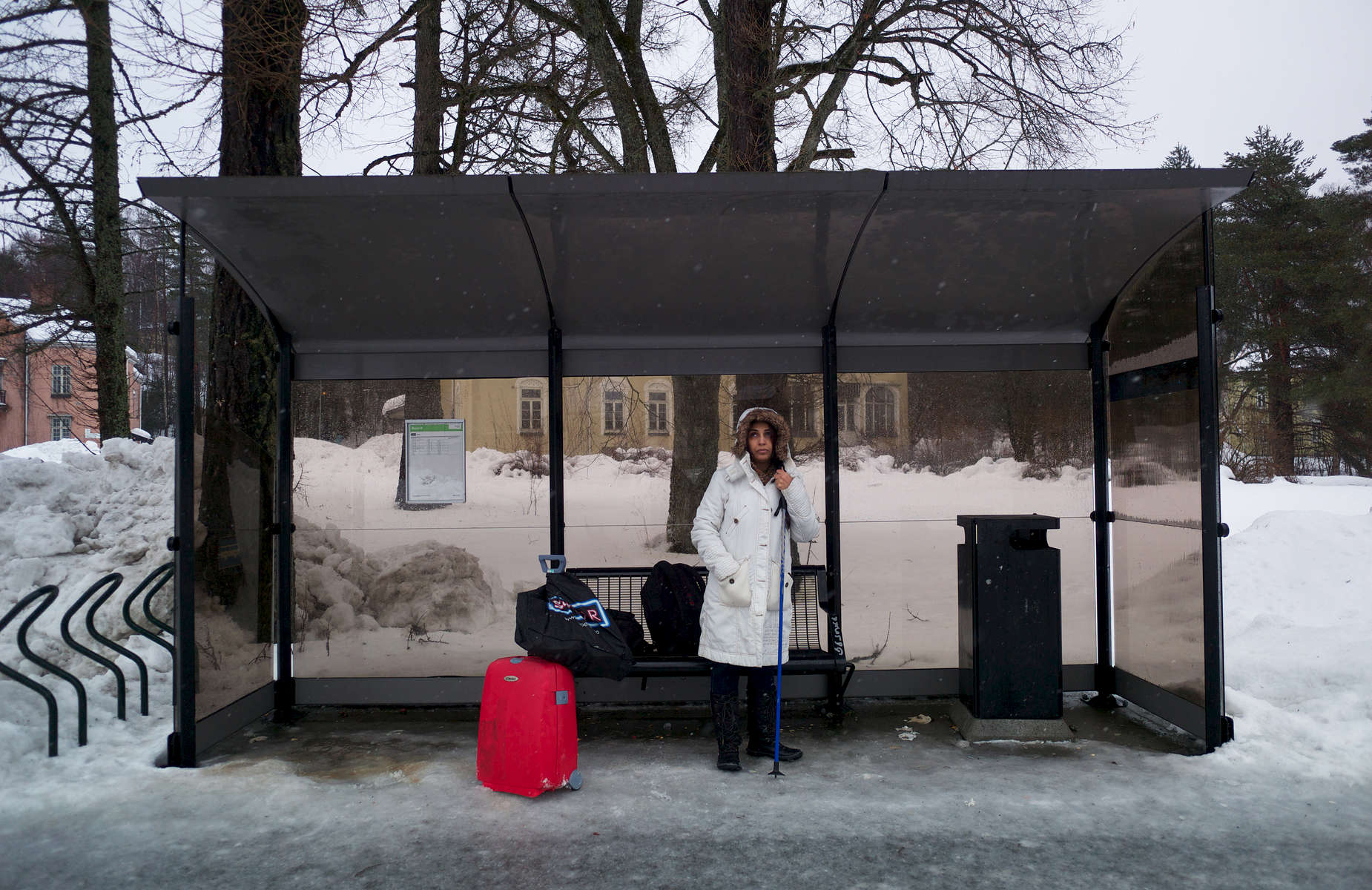 A rejected asylum seeker, with her luggage, stands alone at a bus stop.