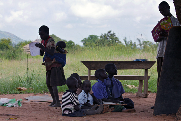 While taking notes a 10-year old girl paces with her one-year old brother on her back, at an outdoor classroom. A teacher stands near a wood board with five students seated on the ground.