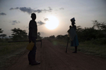 A 12-year old boy and his mother (both holding garden tools) are silhouetted, with the sun raising in the background, as they stand on a dirt road.