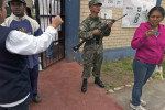 Standing outside an Afro-Peruvian man is directed by a female poll worker on where to cast his vote as a young Peruvian woman stands nearby along with a soldier holding a machine gun.