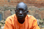 A male Rwandan inmate sits while looking directly into the camera during an interview; he's outdoors with a brick wall serving as a backdrop.