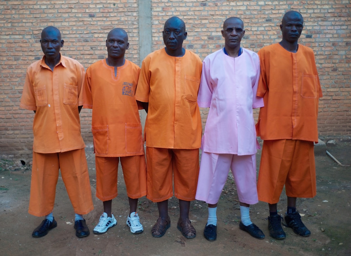Five Rwandan men wearing prison uniforms are standing side-by-side (looking into the camera), outside with a brick wall as a backdrop.