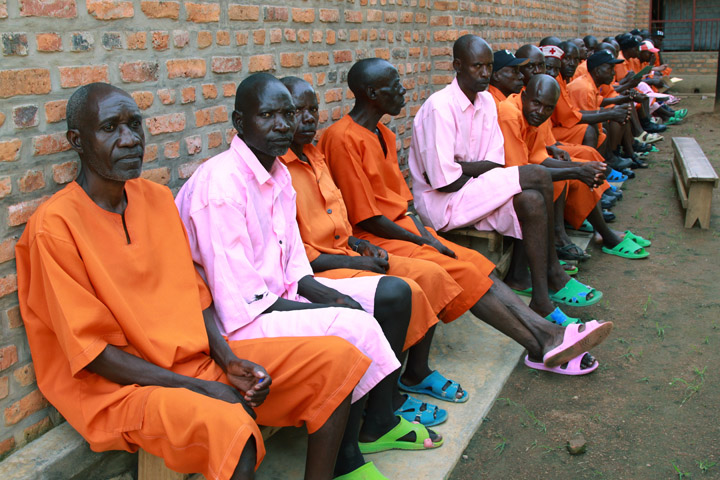 The shot shows a long line of men seated closely together; they're all wearing either an orange or pink prison uniform.