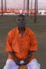 A 58-year old man, wearing a prison uniform, is seated on a chair holding a small piece of paper between his hands. In the distant background are other inmates in prison uniforms.