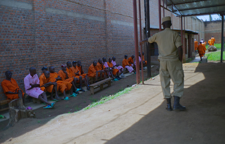 Standing outside a prison guard points and gives commands to several seated inmates; a long line of inmates sit waiting to be interviewed about the atrocities to which they confessed.