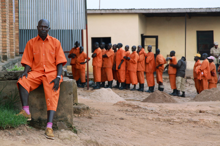 A 45-year old inmate sits outside facing the camera, as 16 fellow inmates are lined up, in the background, and searched as they wait to enter the prison yard - all are wearing orange prison uniforms.