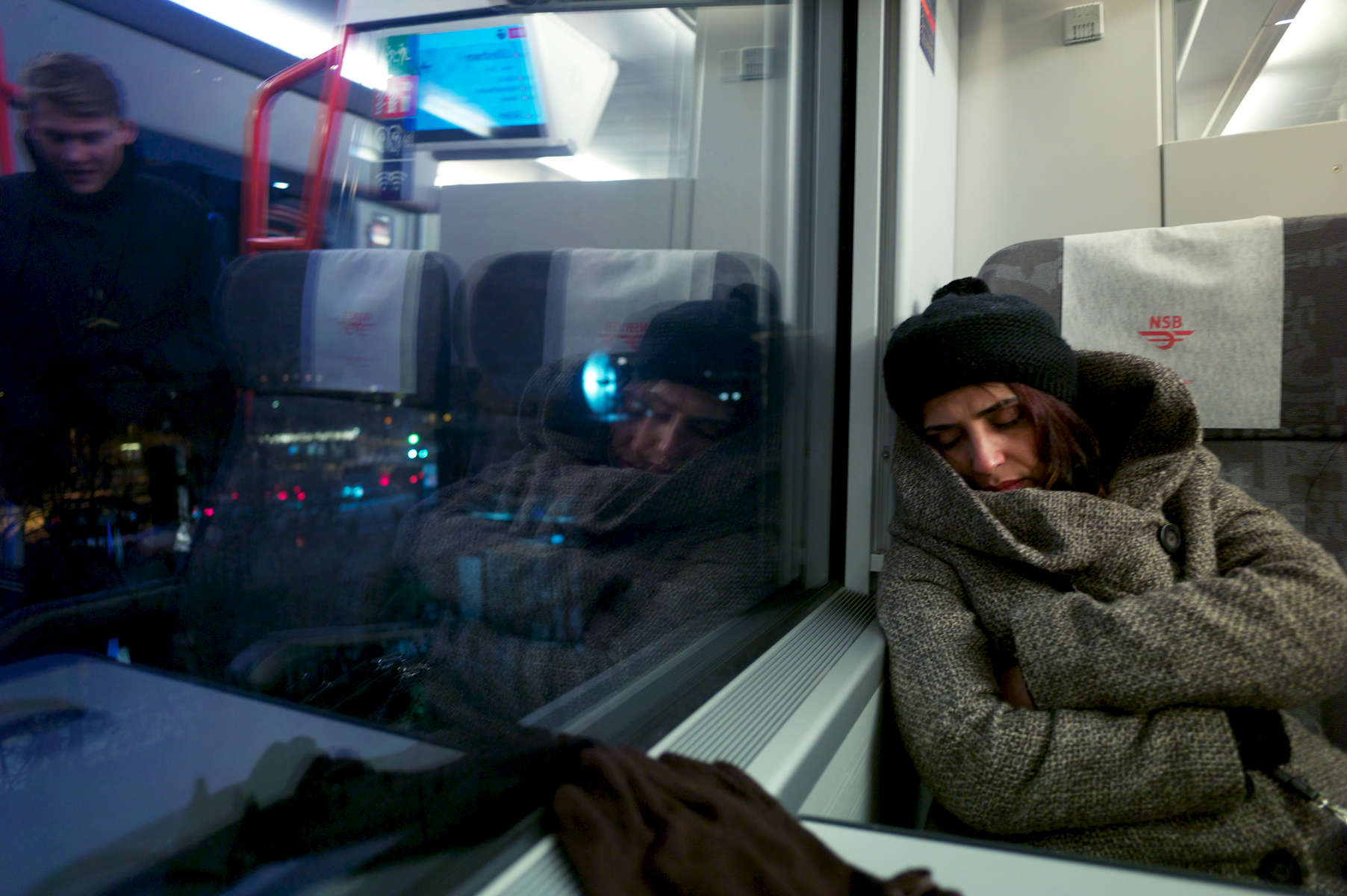 A rejected asylum seeker sleeps on a train as a fellow passenger (who is reflected in the window) approaches her seat.