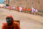 An older Hutu woman sits in a chair outside the walls a prison; there is a group of inmates in the background behind her waiting for visitors.