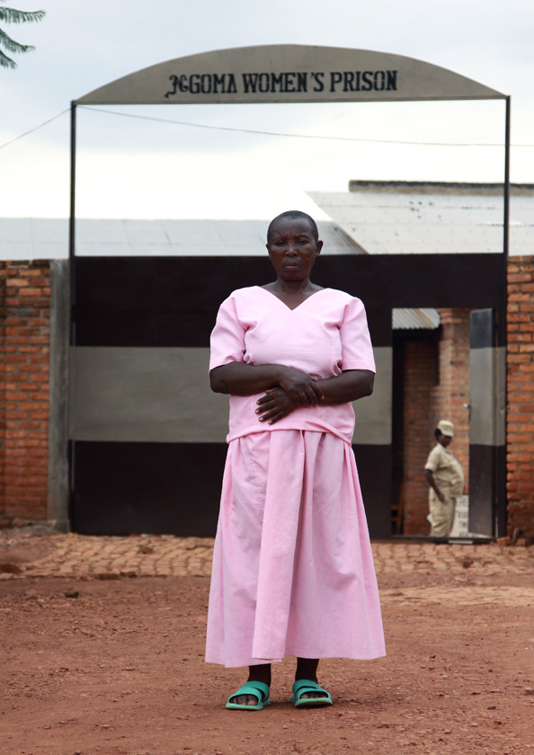 It is a vertical photo of a 64-year old woman, dressed in prison clothing, standing in front of a sign that indicates the name of the prison.