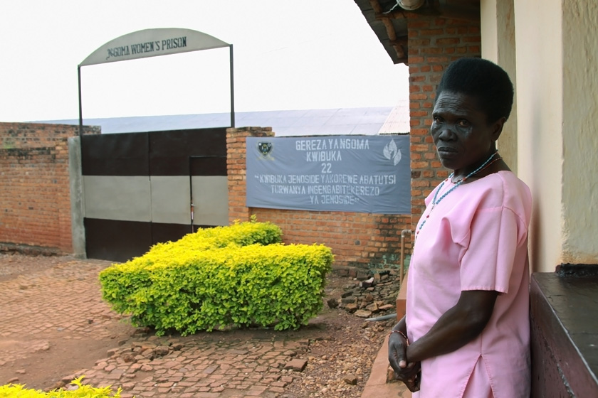 Standing outside a 62-year old woman, dressed in a pink prison uniform, looks into the camera; in the background is a sign with the prison's name.