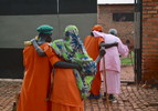 Shown from behind, two elderly Rwandan women are held up by two other elderly women as they walk back to their rooms - all are wearing prison uniforms.