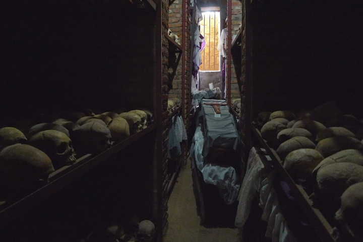 A casket, holding the remains of numerous victims, is displayed in the middle of a very narrow and dark room; the walls on both sides of the room are layered with skulls.