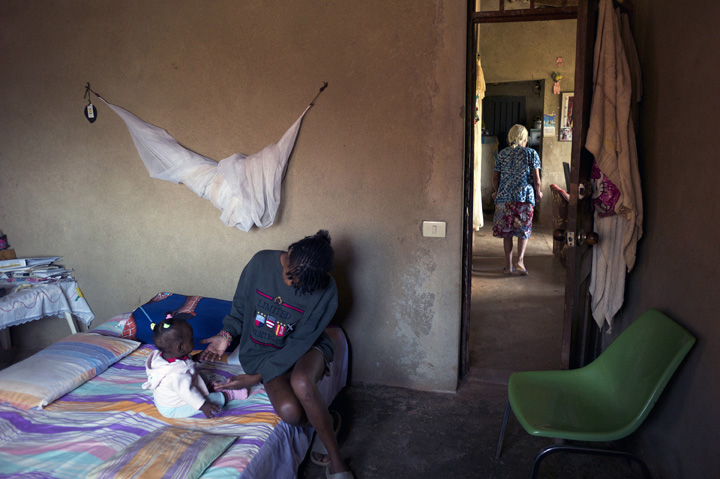 A teenage mother seated on her bed extends her arms for her baby to come; the teenager's grandmother is walking down the hall in the background.