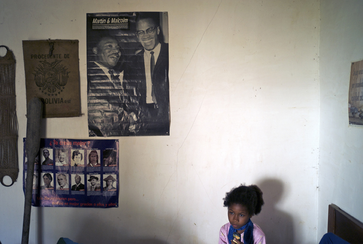 A very young Afro-Bolivian girl is sitting on a bed with her back to a wall that has a poster of Malcolm X and Dr. Martin Luther King, Jr.