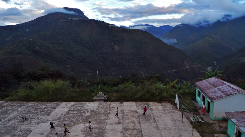 It's a landscape photograph that looks down on a concrete soccer field, surrounded by tropical mountains, as several children kick a ball.