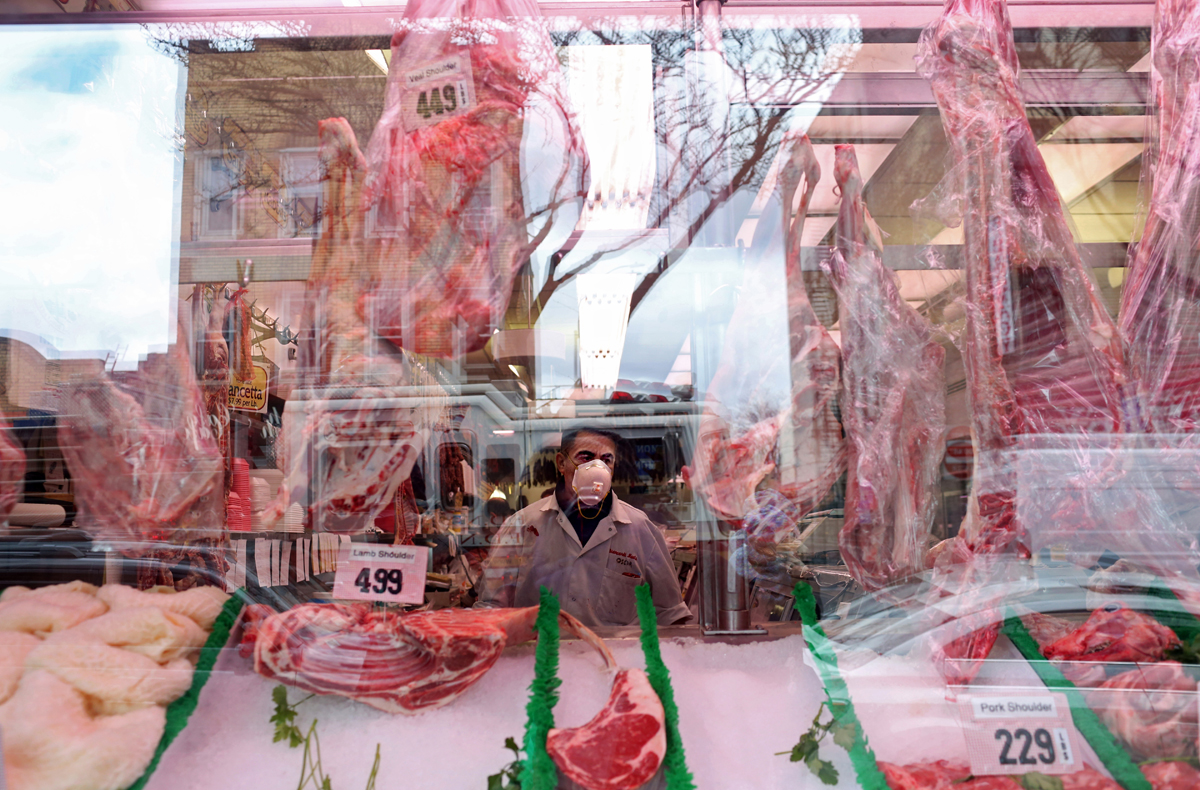 A masked butcher looks out of Biancardi Meats as a line of customers waits to pick up food outside in the Bronx, NY, on April 10, 2020. (Photo by Yana Paskova/For The Washington Post)