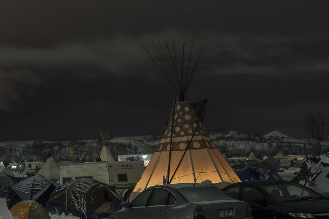 A teepee stands illuminated amidst the parked cars.