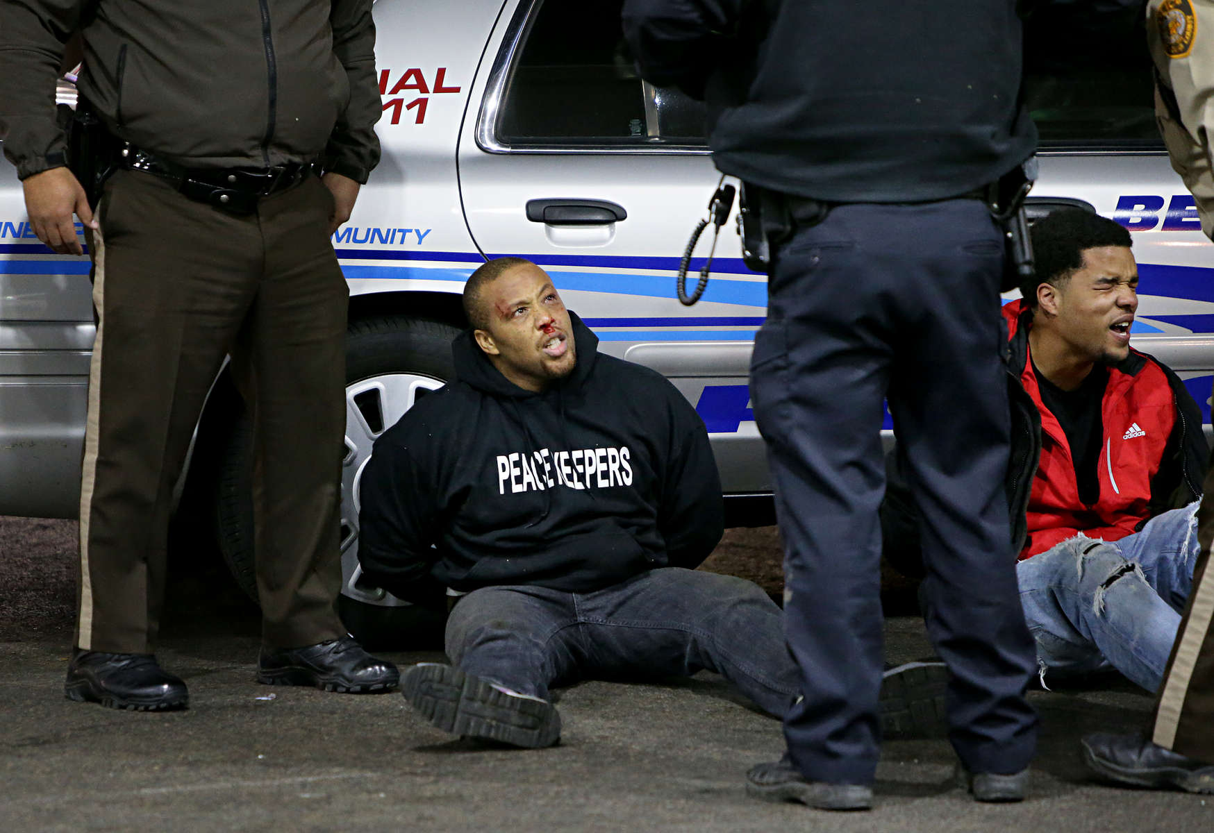 Two men arrested by police during a fight are seated next to a police cruiser on Wednesday, Dec. 24, 2014 on the lot of a gas station where teenager was fatally shot late Tuesday Dec. 23, 2014 at a Mobil gas station on North Hanley Road in Berkeley. The fight broke out after the body of the teen was removed and police and protesters got into a shoving match.Photo By David Carson, dcarson@post-dispatch.com