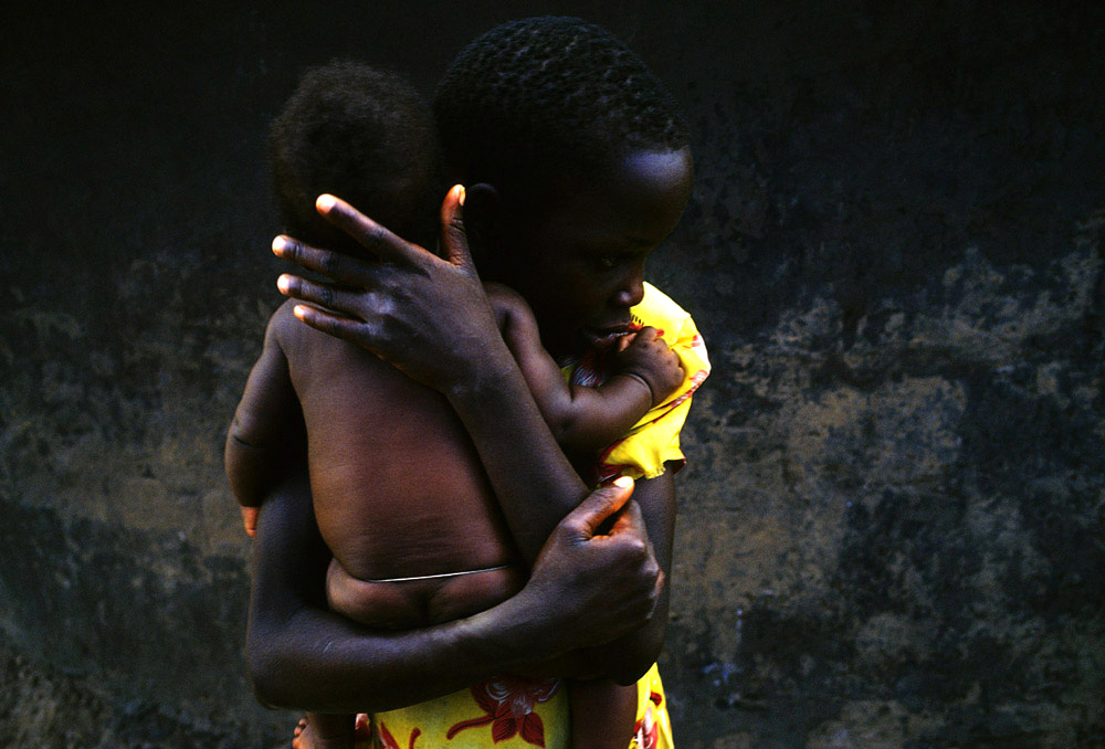 Susan Labol, age 10, hugs her little sister outside her family's hut in Laliya, a rural village in Northern Uganda, 2005. Susan is a night commuter, one of about 20,000 children that sleep in Gulu town, as they are afraid of being abducted by the Lord's Resistance Army (LRA). The rebel group has brought terror to Northern Uganda for almost twenty years, fighting the Ugandan government. The victims are usually children, which are abducted and used as child soldiers and sex slaves. Susan walks 1.5 hours from her home village with her sister Gladys, age 12, every day to sleep at Noah's Arch, an NGO housing children in Gulu. They are too afraid to sleep in the village as an older sister was earlier abducted. 