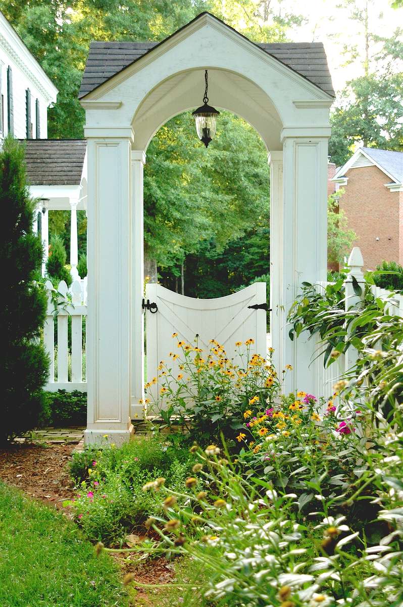 Of timeless design, the white picket fence appears in various interpretations throughout the back yard. Tucked between brick columns, its open picketing gently delineates the property line and includes an arbored gate, inviting neighborly visits. Elsewhere, atop a stone wall and with a scalloped upper pattern, the fence fashionably ensconces a lush lawn beneath the stately grand old oak.