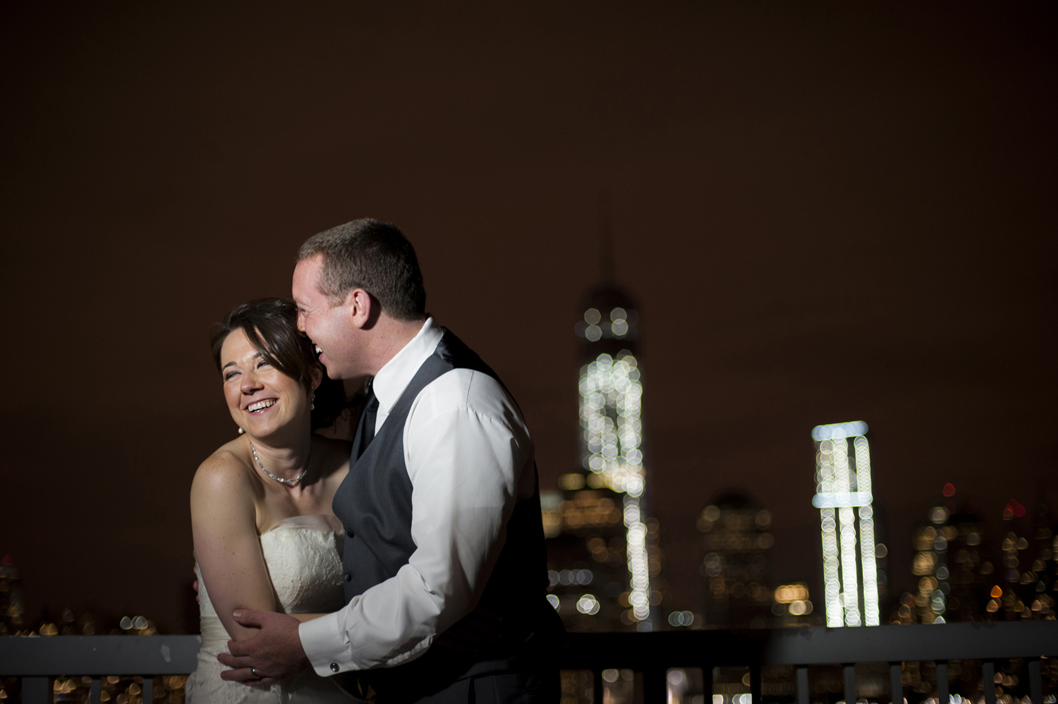 Portrait of bride and groom laughing against the NYC sklyine at night on their wedding day. Jersey City wedding photographers
