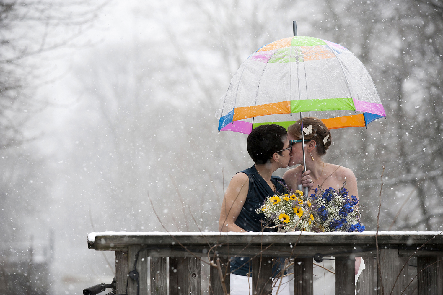 Brides kissing in the snow under colorful umbrella during their winter wedding at Lambertville House. Lesbian wedding