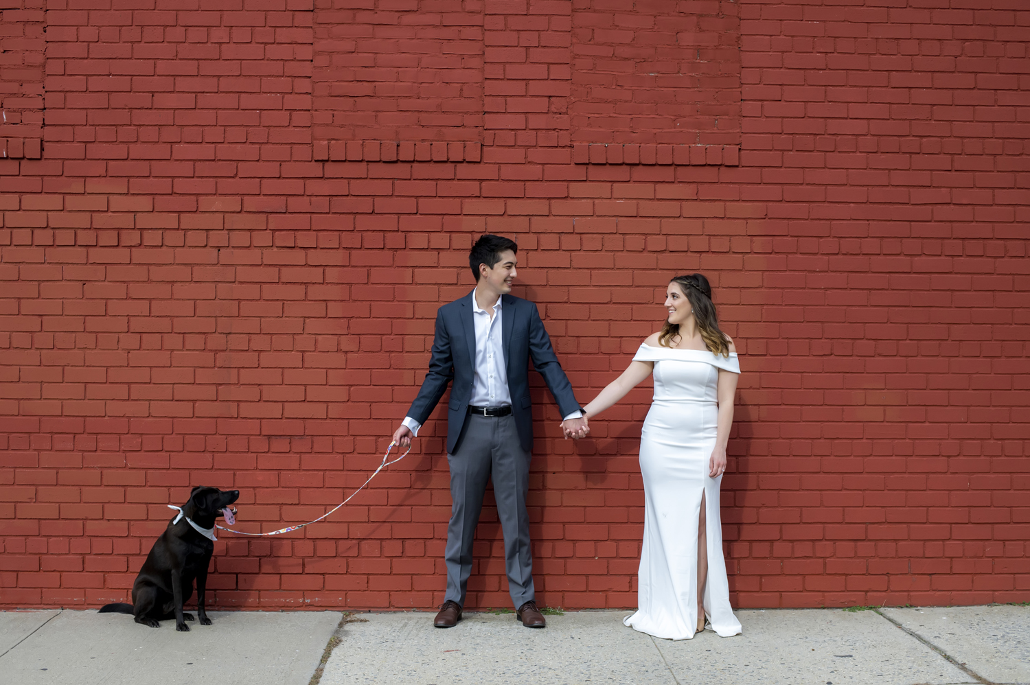 bride and groom and their dog pose for a portrait against a red wall on their wedding day in Astoria, Queens. Queens wedding photographer