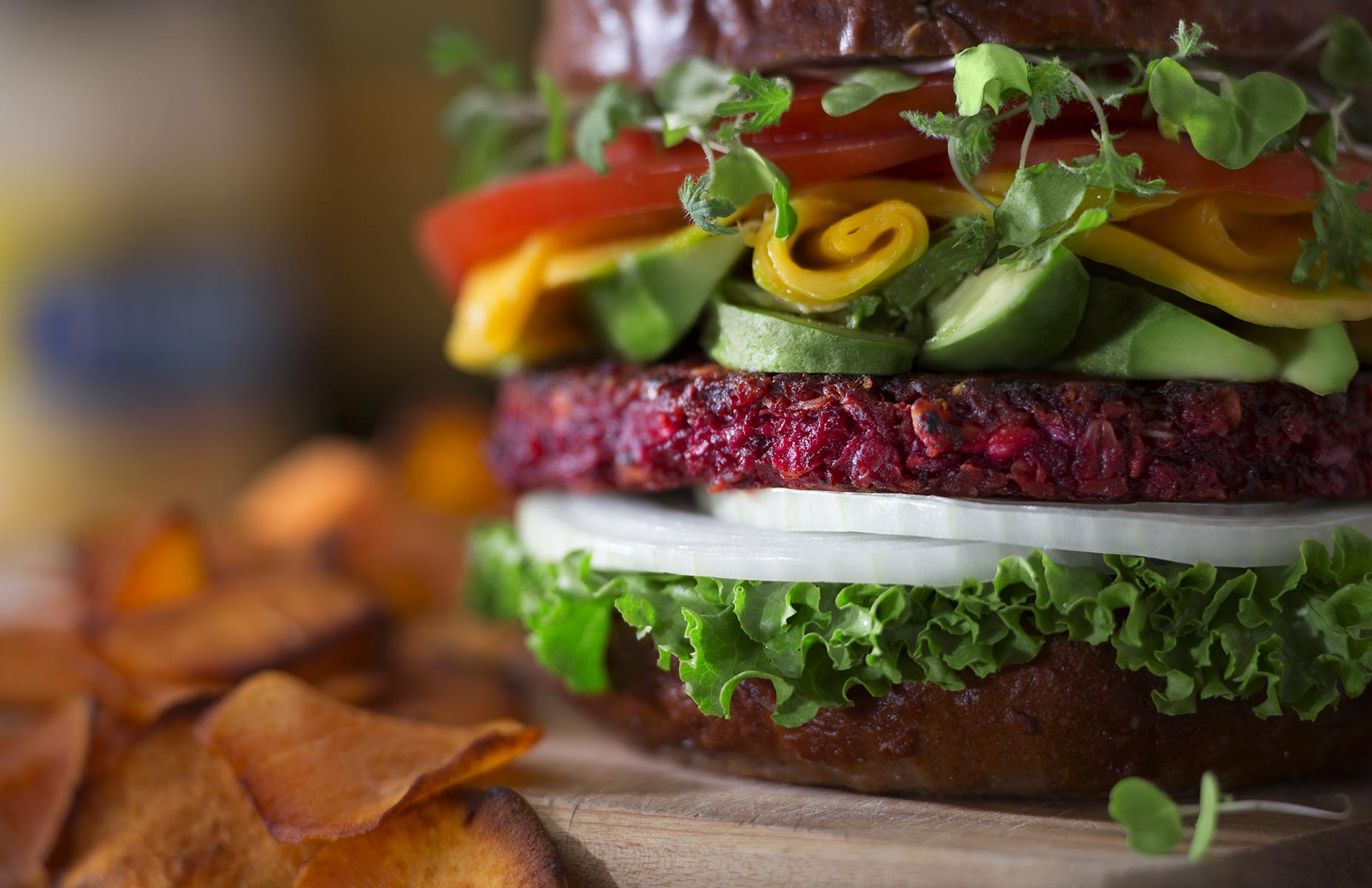 A low angle view of a beet patty hero sandwich made with raw white onion and tomato slices, rolled mango slices substitue for cheese and avacado chunks stacked high on a pretzel bun. Lettuce and bean sprouts finish this entree which sits on a distressed wood cutting board and is served with roasted sweet potato rounds. Dramatically lit from the left, the background is muted and out of focus exhibiting strong bokeh effect cropped in a landscape orientation.