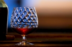 Camera is at 0º and the lighting is frome back and right of frame in this landscape aspect product and beverage image. The single snifter is partially filled with a hint of the bottle on the left side and a cork stopper is placed between all composed on a weathered wood bar top. The soft focus background exhibits a desireable bokeh effect in its rich brown and blue hues.