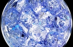 The camera angle is from directly above and the light is from below in this fast flash duration and close-up photograph of ice and soda in cyan and blue hues against black. Bubbles of efforvesence stream to the fluid surface almost engulfing while surrounding the smallish clear ice cubes. Presented at 11{quote}x17{quote} plus bleed.