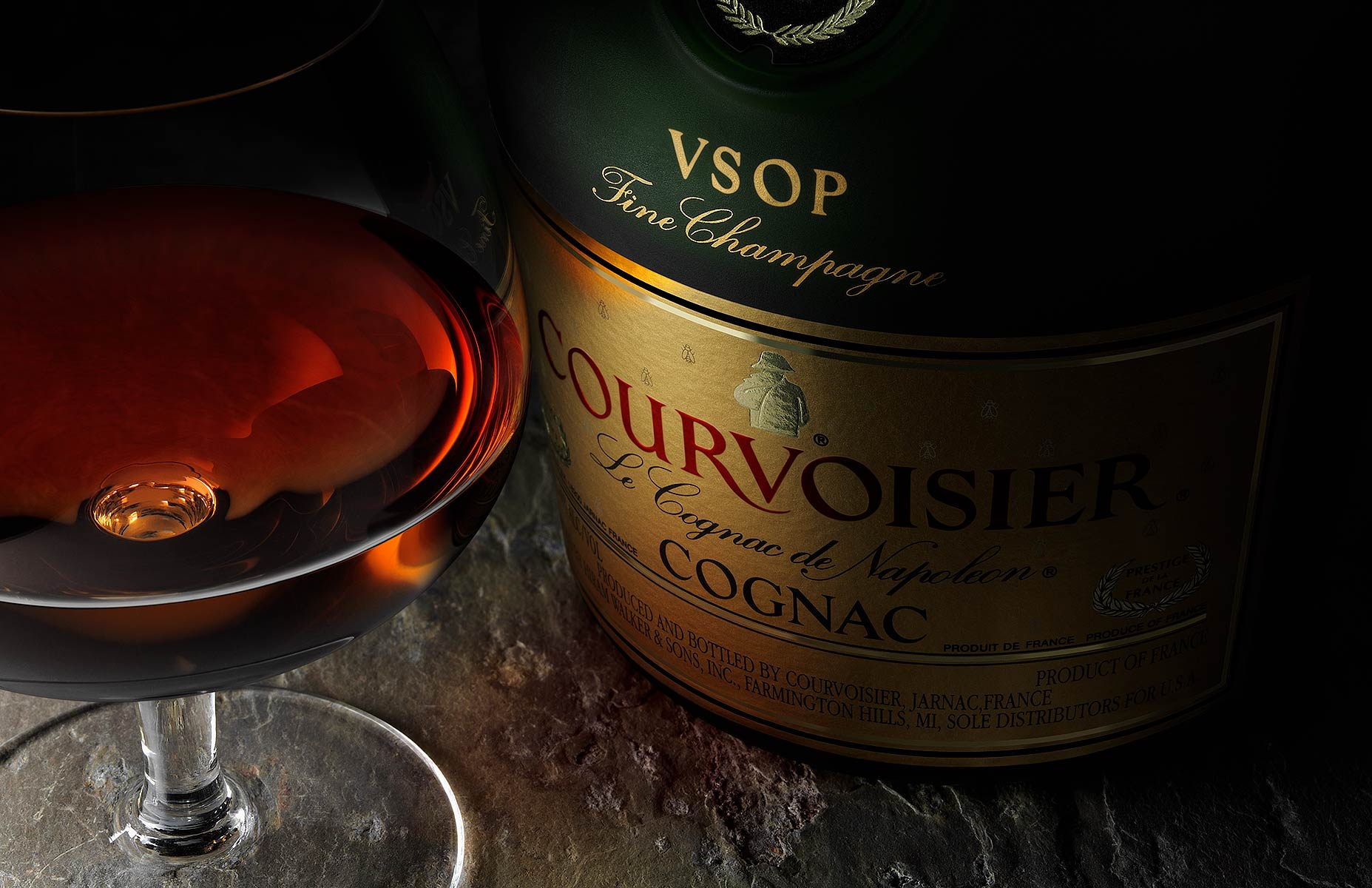 A bottle of VSOP Courvoisier dominates the horizontal frame with a partially filled snifter. The camera angle is at 45º and the subjests sit tightly on a rough hewn stone bar top. The feeling is moody and the soft light originates on the left side highlighting the glasss and label. A reddish glow also illuminates the lower label from light passing through the fluid.