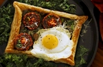 Fried Egg En Croute is an entree made with Phyllo dough shaped into a rectangular shell photographed from above. It is filled with asparagus, roasted tomatos, sauteed garlic, parmesan cheese then baked with spices and herbs. The delectable treat is finished with a fried egg and served on a bed of kale leaves all resting on a cast iron grill pan on dark wood. The image is in a landscape orientation and softly back lit.
