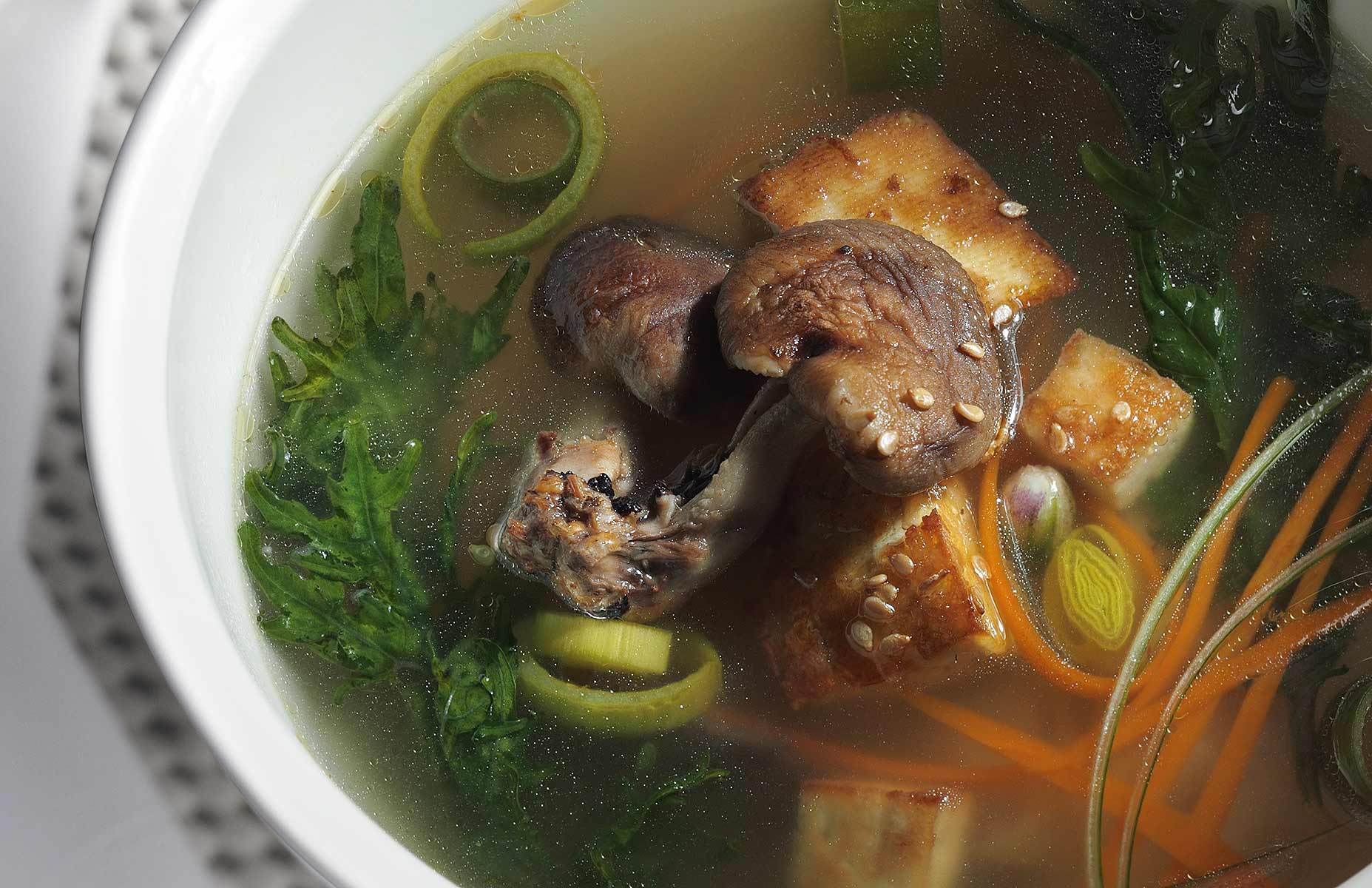 This image of Miso Soup is photographed at a 45º angle and is cropped tightly in a landscape orientation. The dish incorporates leeks, carrots, seaweed and shitake mushrooms in a miso broth with sprinkles of sesame seeds in a white bowl. The back and side lighting technique creates depth in the broth and a sculptural effect to the mushrooms.