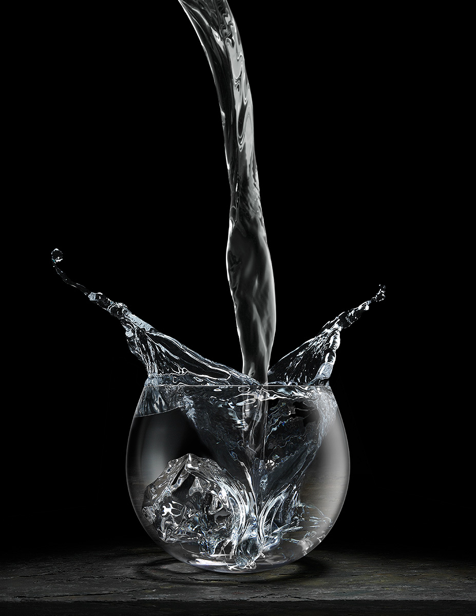 Pouring Vodka Splash is an example of high speed photography capturing the excitement of a hard pour into a cocktail glass creating splashes. The glass is sitting on a dark slate surface with a black background. This image is in portrait orientation. The camera is dead on and the soft and dramatic light source is from the right. The image is monochromatic with strong highlights in the glass, splash and pouring fluid.