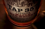Product image exhibiting forced perspective dominates the visual appeal of the still life. The lighting is focused on the label of the bottle with an internal glow from the interior fluid. A cork rests to the right of the maple infused rye whiskey bottle and has a rim light effect. The subject is in sharp focus top to bottom and all sit on a rough hewn wood plane. The image is cropped in a landscape format.