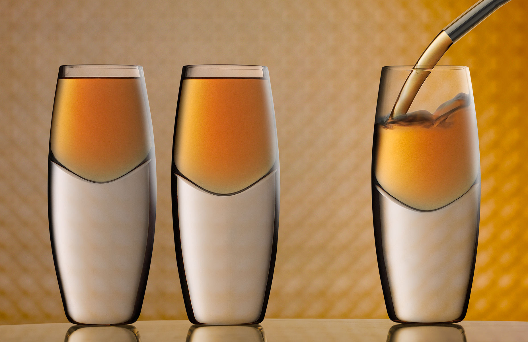 Three shots, the two on the left are already full with an aperitif pouring into the glass on the right side of frame. The background exhibits beautiful bokeh effects in the predominantly orange hue. The glasses are composed on a slightly reflective white with black and grey veined marble surface. The image is cropped horizontally and the camera is dead-on to the subject.