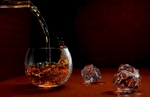 Whiskey pouring from a bottle into a round lowball glass on a mahogany wood surface which fades to black with liquid splashing about. This example of high speed photography exhibits incredible detail while two ice cubes melt on the surface. This picture is in landscape orientation with a low angle of view and strong light source from the right.