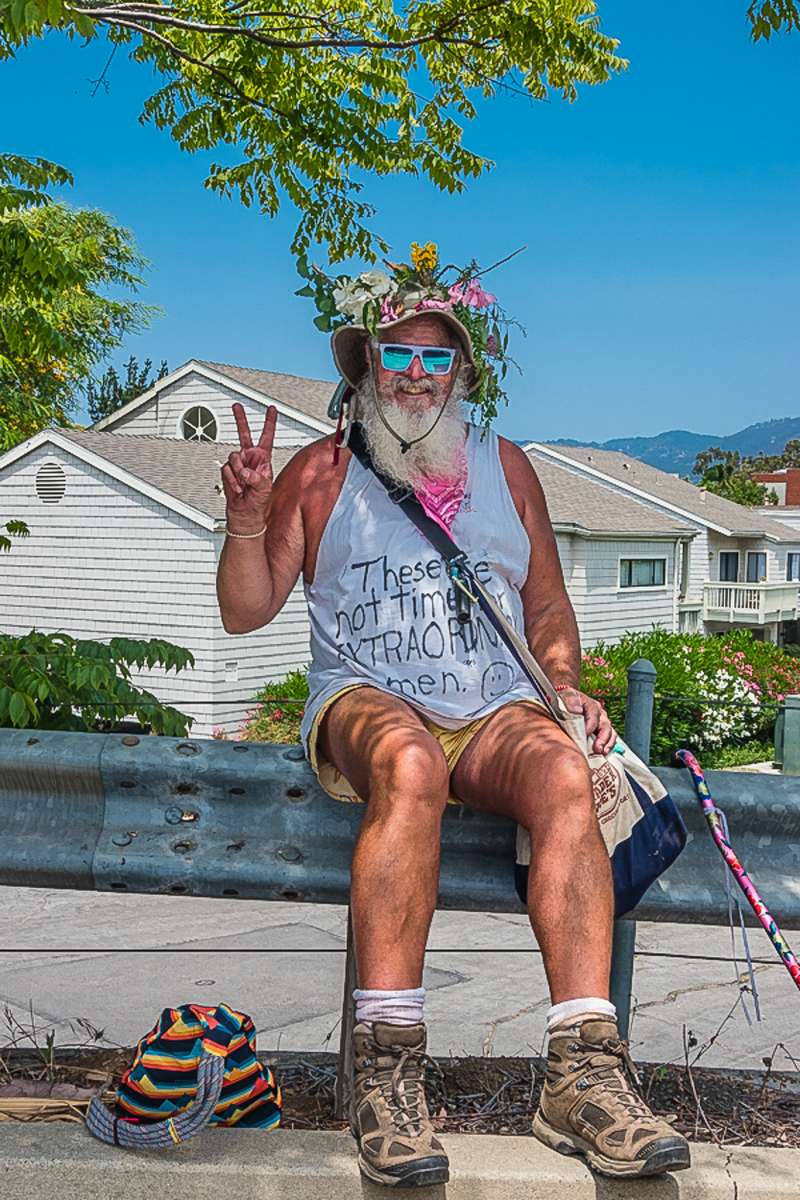 Funny man in flowered hat and reflective sunglasses wearing a cryptic t-shirt while sitting on a guardrail in Santa Barbara, California.
