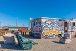 A graffiti laden trailer with the word sore painted on it along with symbols and several lounge chairs ouside in the desert sand in front of it.