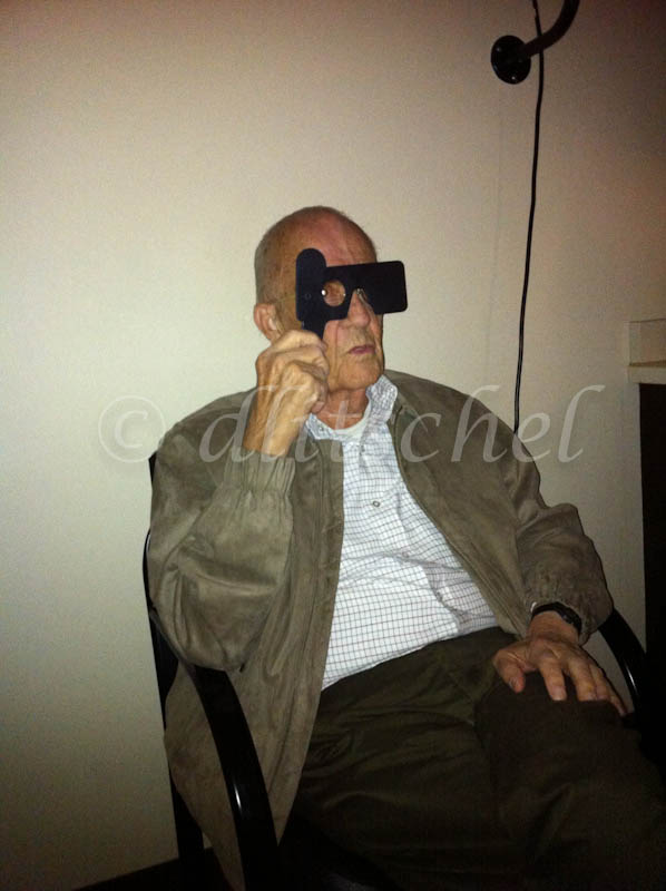 A senior citizen sits in eye exam holding a device that allows him to see through one eye only while looking at the eye chart.