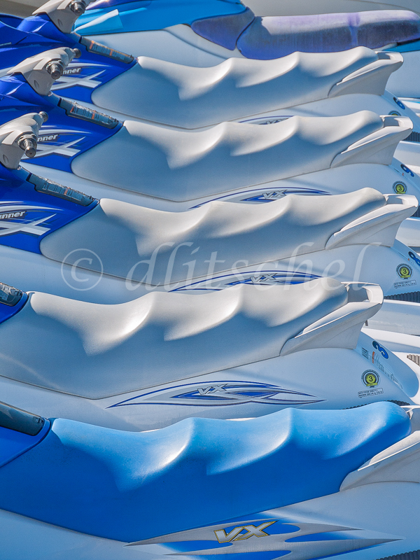 The vertical line up of See-Doo water craft with white undulating seats appears to look like either a row of white cap waves in the ocean or whipped cream topping. The seats provide graphic and abstract pattern of repeated form. Some handle bars are visible and the {quote}VX{quote} model designation appears on four of the vehicles.