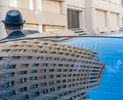 A limousine driver, in a black hat, stands facing away whose head and neck are visible because he is blocked by the rear window of his limo. The window reflects the high-rise buildings on Powell St. in San Francisco.
