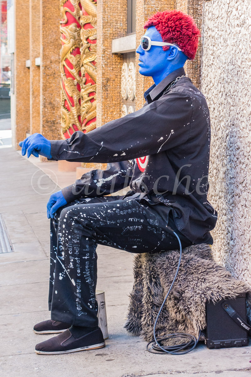 A young adult male street performer with a red wig and blue body paint on his face, neck and hands, dressed in black and sitting on his amplifier ready to perform for money in Chinatown, San Francisco, California.