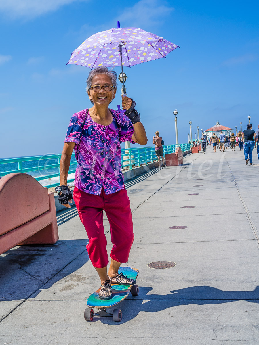An Asian grandmother smiles as she holds a parasol and skates on her skateboard on the pier in Manhattan Beach, California.