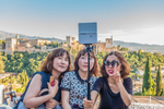 Three Asian females sit, posing together, at the Mirador San Nicolás, for a group selfie with one gesturing with the {quote}V{quote} sign with her fingers and with the Alhambra in the background in  Granada, Spain