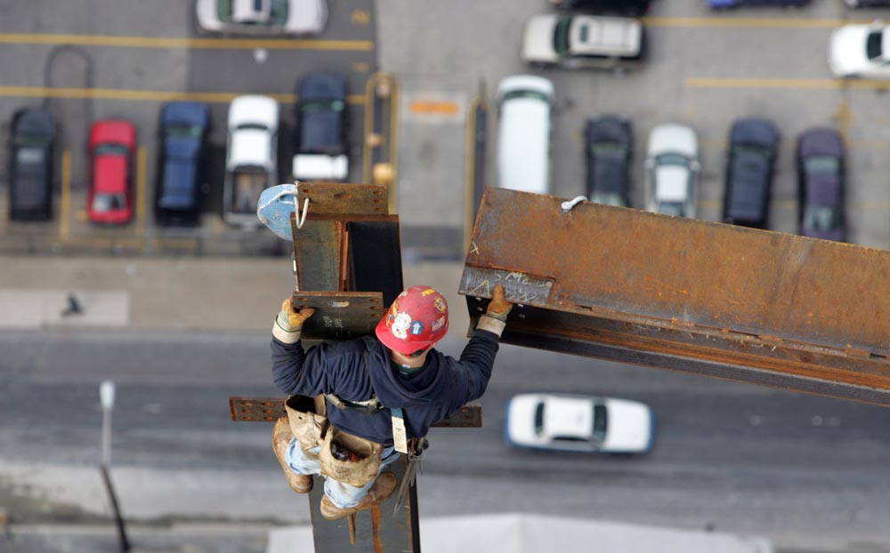 A third-year apprentice, guides a steel beam into place on the 43rd floor. He is tied off on a column, approximately 638 feet above 18th Street, where a motorist passes.