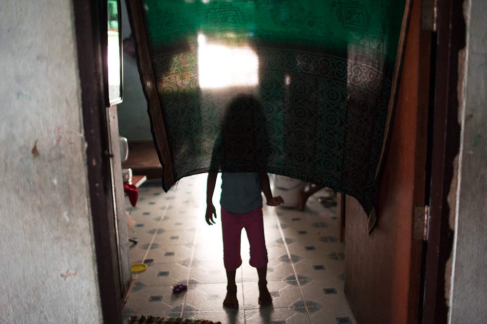 A young asylum-seeker stands behind a curtain at the entrance of a one room apartment inside a low income building on the outskirts of Bangkok. Mar. 2015