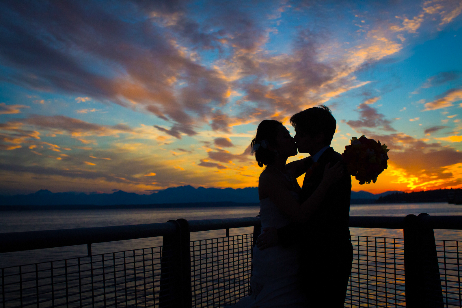 The bride & groom are silhouetted as they kiss in front of a beautiful sunset on the Seattle waterfront during their wedding at the Seattle Aquarium. (Wedding Photography by Scott Eklund - Red Box Pictures)