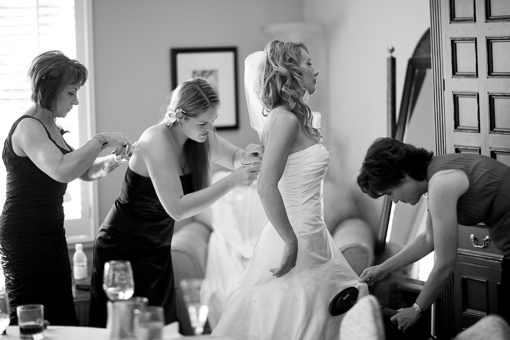 Bride Pauline gets help getting ready from her mom and a bridesmaid as the wedding coordinator steams the wrinkles out of the wedding dress before the start of the wedding at the Golf Club at Newcastle near Seattle. (Wedding Photography by Scott Eklund/Red Box Pictures)